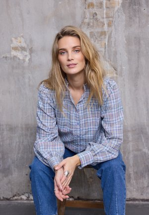 Chemise à carreaux bleu et blanc avec manches bouffantes, portée avec un jean en denim bleu. Mains posées sur les genoux, bague en argent à la main gauche.