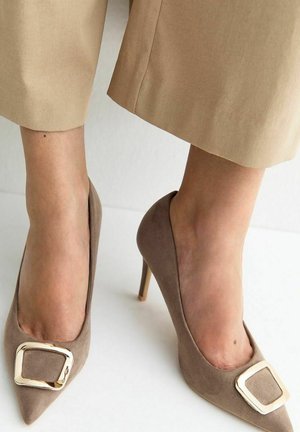 Voeten met beige suède hoge hakken met grote gouden vierkante gespen, gecombineerd met lichtbruine cropped broeken tegen een witte achtergrond.