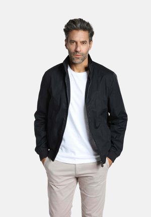 Blouson aviateur noir avec une fermeture éclair à l'avant et un col montant, associé à un T-shirt blanc à col rond. Un pantalon de couleur claire complète la tenue.