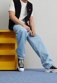 Gilet in pile nero su una maglietta bianca, jeans larghi azzurri chiari e sneakers nere con suole bianche, seduto su un comò giallo.