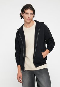 Melna zibspējas džemperis ar kapuci, ar ribotām aprocēm un priekšējiem kabatām. Nēsāts virs gaiši brūna tee krekla un apvienots ar pelēkiem džinsiem.