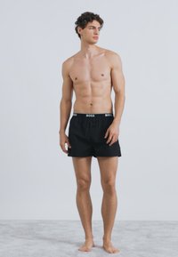 Čierne boxerky z bavlny s elastickým pásom so značkou "BOSS" a voľným strihom. Model má štíhlu postavu.