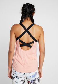 Jasnoróżowy sportowy top na ramiączkach z czarnym wzorem na krzyż na plecach, z odkrytymi plecami i gładką teksturą. Połączony z wzorzystymi legginsami.