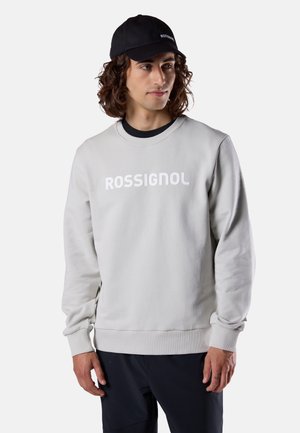 Mann mit lockigem Haar, der einen hellgrauen Rossignol-Sweatshirt, schwarze Hose und eine schwarze Kappe trägt und seitlich auf weißem Hintergrund schaut.