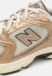 Beige och krämfärgad sneaker med nätpaneler, läderdetaljer och en texturerad sula. Har vita snören och en mörk "N"-logotyp.