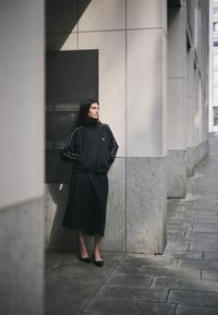 Chaqueta deportiva negra con detalles blancos, combinada con una falda negra. Tejido suave, ajuste relajado, posando frente a un muro de piedra.