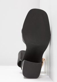 Chaussure à talons hauts noire avec une tige en cuir lisse, un bout arrondi et une semelle en caoutchouc texturée. Présente un accent métallique.