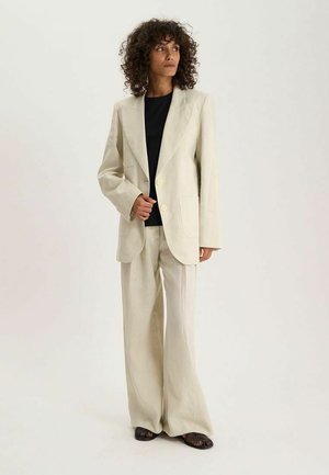 Personne aux cheveux bouclés portant un costume beige oversize avec un pantalon à jambes larges et une chemise noire, debout devant un fond clair uni.