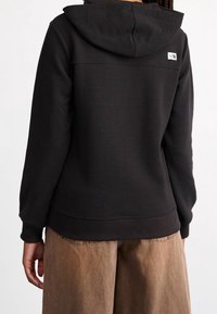 Felpa nera con cappuccio e texture a coste, con un piccolo logo sulla parte superiore della schiena e un orlo a contrasto. Abbinata a pantaloni marrone chiaro.