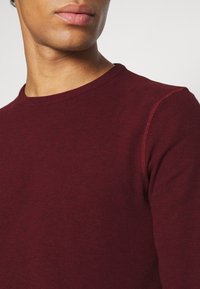 Bordowy sweter z okrągłym dekoltem i prążkowaną fakturą. Zbliżenie pokazuje detal szwu na ramieniu oraz gładkie wykończenie materiału.