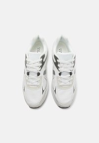 Valkoiset urheilujalkineet, joissa on hengittävä verkkopinta, harmaat yksityiskohdat, pehmustettu sisäpohja ja kohokuvioitu kumipohja.