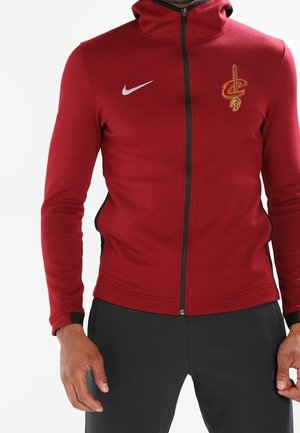 Tummanpunainen vetoketjulla varustettu huppari, jossa Cleveland Cavaliersin logo ja valkoinen Nike-swoosh, miehen päällä mustien housujen kanssa.