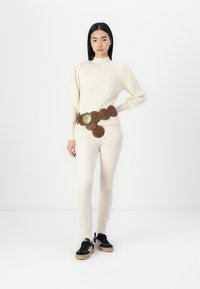Pulover din tricot crem cu mâneci bufante, pantaloni bej ajustați și o curea maro circulară cu accente metalice, purtată cu adidași negri.