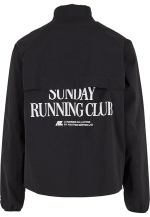 Musta takki, jossa on valkoiset tekstit "Sunday Running Club" ja pienempi teksti "A Running Collective by Another Cotton Lab" selässä.