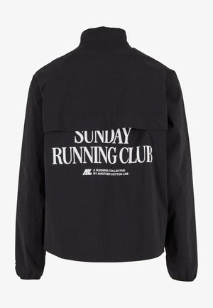 Chaqueta negra con texto blanco "Sunday Running Club" y un texto más pequeño "A Running Collective by Another Cotton Lab" en la parte de atrás.