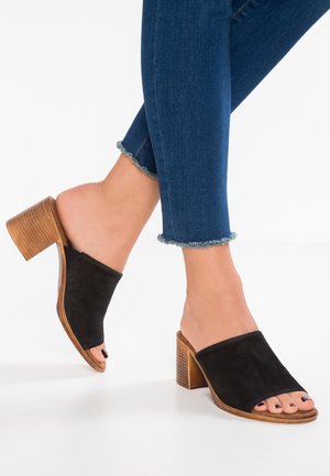 Mustat mokkasaappaat, joissa on leveä, litteä hihna ja avoin kärki. Palikkakorko on teksturoitua ruskeaa puuta, tarjoten vakaata tukea.