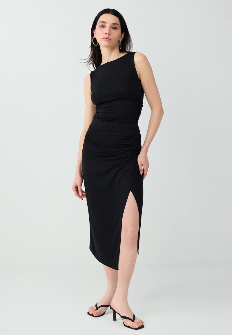 Femme en robe midi noire sans manches avec fente latérale, portant des créoles et des sandales à talons noires, debout devant un fond uni.