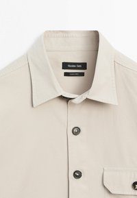 Beige Hemd mit Knöpfen, spitzem Kragen, schwarzen Knöpfen, Brusttasche und sichtbarem Massimo Dutti Markenetikett im Kragen.