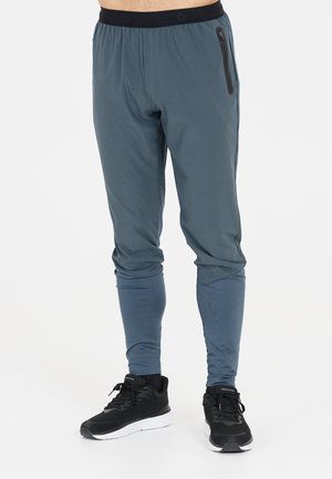 Mann trägt blaue Slim-Fit-Jogginghosen mit einer Reißverschlusstasche auf der rechten Seite und schwarze Laufschuhe mit weißen Sohlen.