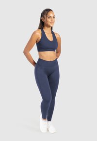 Marinblå sport-bh och högmidjade leggings i texturerad tyg, i kombination med vita träningsskor. Modellen står med händerna bakom ryggen.