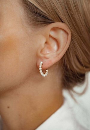 Boucle d'oreille en or ornée de petites perles blanches, présentant un design courbé. Gros plan de la boucle d'oreille sur un fond de peau neutre.