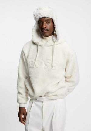 White fleece hoodie with embossed "BOSS" logo, paired with a matching white fur-lined hat. Model stands with hands behind back, wearing light pants.