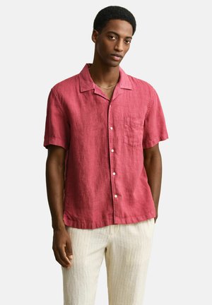 Man draagt een rood overhemd met korte mouwen en lichtbeige broek met strepen, staand voor een effen witte achtergrond.