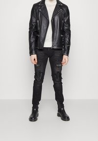 Chaqueta de motociclista de piel negra con remaches plateados, suéter blanco ajustado, jeans negros desgastados con acentos de pedrería y botas negras hasta el tobillo.