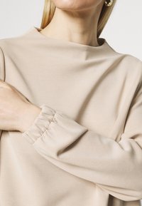 Beżowa bluzka z wysokim dekoltem, miękkiego materiału i zgromadzonymi mankietami. Gładka tekstura z subtelnym marszczeniem, podkreślającym pozycję ramion.
