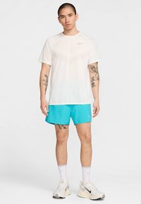 Weißes sportliches T-Shirt mit strukturiertem Muster und kurzen Ärmeln, kombiniert mit leuchtend türkisfarbenen Shorts; Modell trägt weiße Socken und graue Turnschuhe.