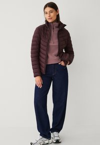 Giacca puffer corta di un viola scuro con trapuntatura a rombi, colletto alto e zip frontale, indossata sopra un pullover viola chiaro e jeans larghi di colore scuro.