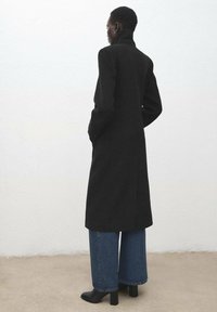 Długi czarny płaszcz z wełny, z wysokim kołnierzem i bocznymi kieszeniami. Luźne dżinsy i czarne botki na słupku. Minimalistyczny design.