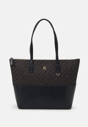 Black tote bag with leather bottom, geometric patterned upper, gold logo and ring detail, and two long shoulder straps with zipper closure.