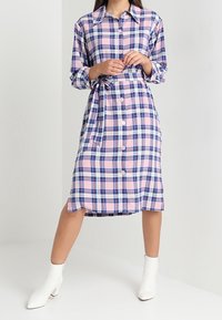 Kvinna som bär en rosa och blå rutig midi-klänning med knappar och midjeband, tillsammans med vita ankelboots, stående med korsade ben.