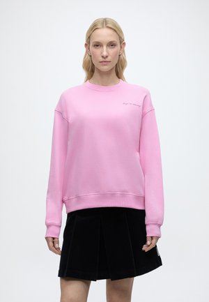 Blonde Frau trägt ein pinkes Sweatshirt mit dem Schriftzug "keep on moving" und einen schwarzen Faltenrock, steht vor einem schlichten weißen Hintergrund.