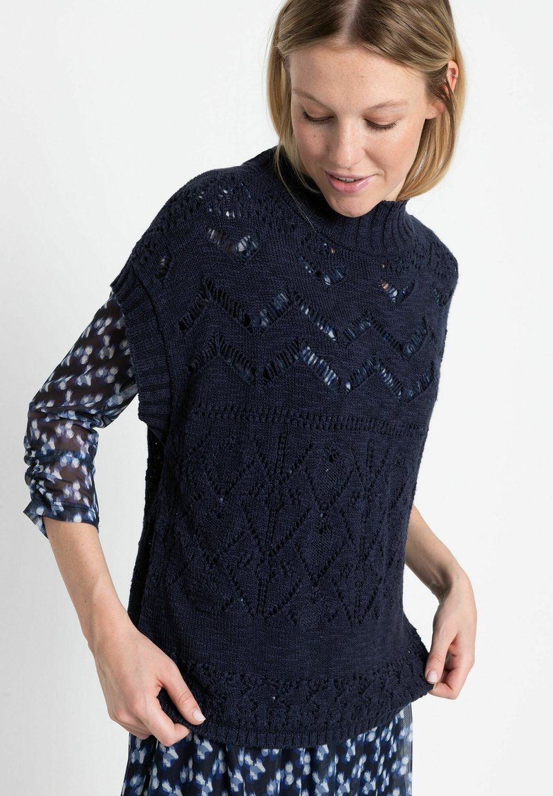Pull en maille bleu marine avec des motifs texturés et des tissages ouverts, doté d'un col haut et d'emmanchures larges, porté par-dessus une chemise à motifs.