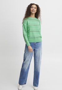 Pull en maille vert clair avec des rayures ajourées, associé à un jean en denim bleu délavé et ample, ainsi qu'à des chaussures blanches à bout pointu.