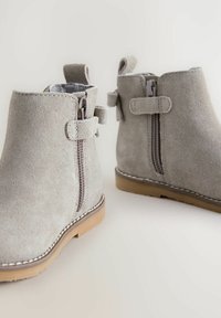 Stivaletti alla caviglia in suede grigio con cerniere laterali, dettagli con linguetta e suole in gomma marrone chiaro con cuciture a contrasto lungo i bordi.