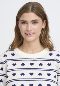 Pull en maille crème avec rayures horizontales bleu marine et motifs en forme de cœur. Encolure ronde côtelée. Texture douce et coupe décontractée.