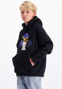 Zwarte hoodie met een graphic van een beer met een pet, grote frontzak en geribbelde boorden, gecombineerd met lichtblauwe jeans.
