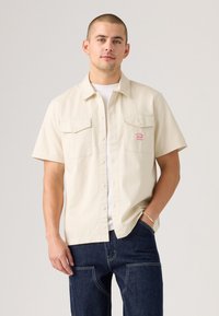 Short-sleeve, cream-colored button-up shirt with two chest pockets and a small branded label, made from a lightweight fabric.