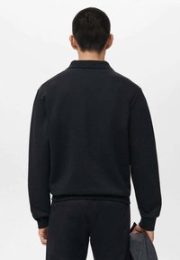 Suéter negro de material suave, con cuello y mangas largas. La vista trasera muestra un corte relajado y dobladillo acanalado, sosteniendo una chaqueta gris en la mano.