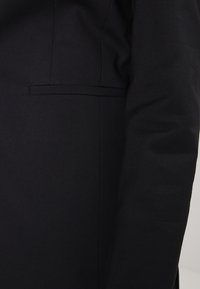 Svart skräddarsydd kavaj med en elegant design, slät yta och en liten sidoficka. Rena linjer och väldefinierad silhuett.