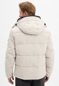 Witte gewatteerde jas met een capuchon, voorzien van een bruine nepbont voering bij de kraag, een rits aan de voorkant en zwarte hardware accenten.