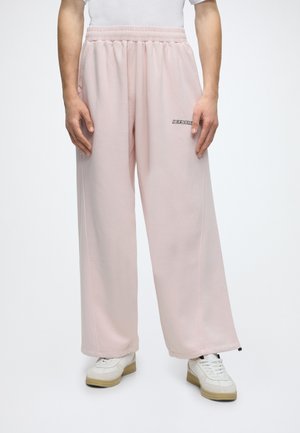 Personne portant un pantalon de jogging ample rose clair avec une taille élastique et des baskets blanches, debout devant un fond blanc.