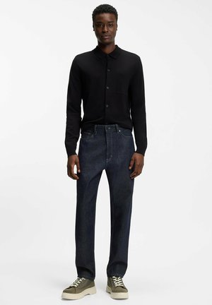 Chemise noire boutonnée avec une poche poitrine, associée à un jean bleu foncé et des baskets montantes vert olive avec des semelles et des lacets blancs.