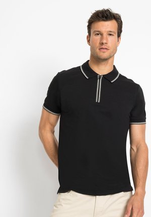Polo noir avec un col zippé, des manches courtes et des accentuations blanches rayées au niveau du col et des manches. Matière en coton lisse.