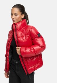 Giacca puffer rossa con colletto alto, finitura lucida, frontale con zip e una toppa sulla manica sinistra; presenta una silhouette squadrata e tasche laterali.