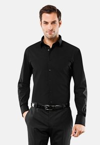 Camicia nera a maniche lunghe con colletto strutturato, realizzata in tessuto liscio, indossata con pantaloni neri e una cintura.