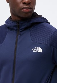 Άνδρας φοράει μπλε μπουφάν The North Face με φερμουάρ, με κεντημένα πάνελς στους ώμους και κουκούλα.